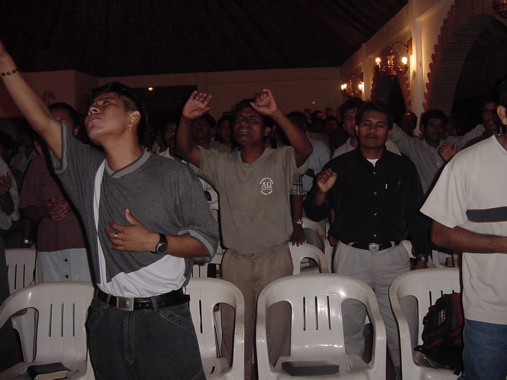 A group of people stand with raised hands during a worship service in a warmly lit building, with musicians and leaders participating in the background.