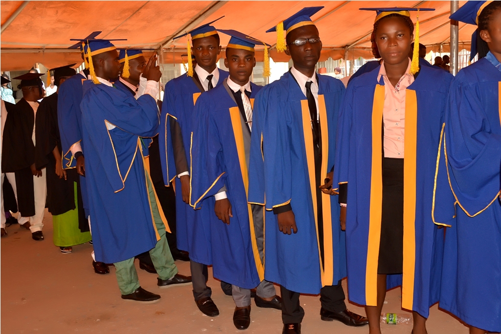 Students wearing blue and yellow graduation gowns and caps stand in a line under a tent during a commencement ceremony.
