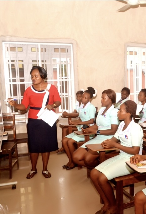 A woman stands at the front of a classroom using a whiteboard to teach nursing students dressed in light green uniforms seated at desks.