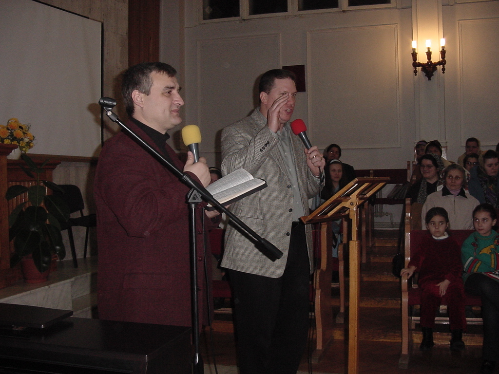 Pastor David and Dan stand at the front of a church, each holding a microphone, speaking to a congregation seated in pews.