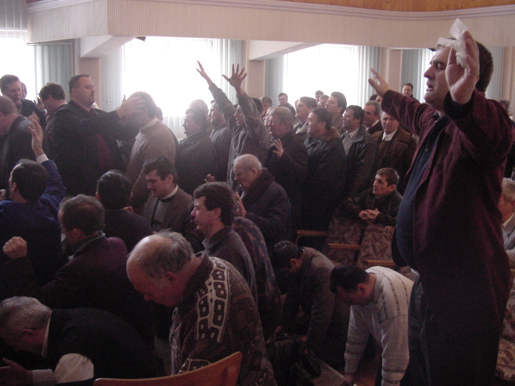 A large group of men pray together in a church sanctuary, many raising their hands or laying hands on others in prayer- alter call