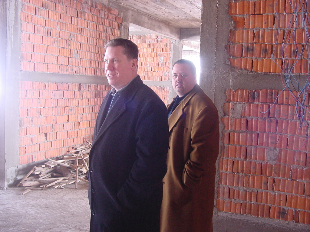 Pastor David and friendin long coats stand inside an unfinished brick building, observing the area.