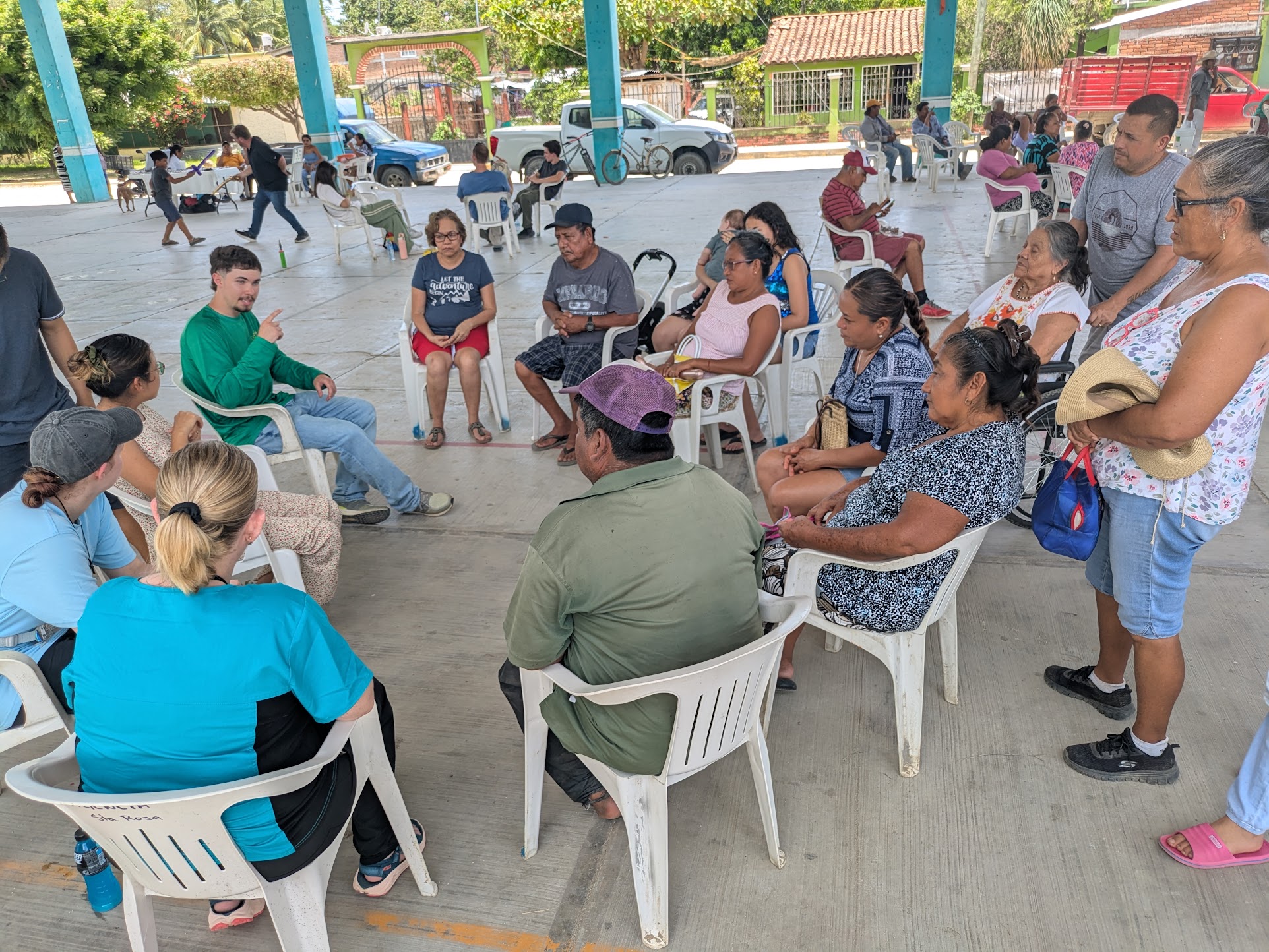 A group of people sit in a circle of white plastic chairs under an open-air pavilion, engaging in conversation during a community event.