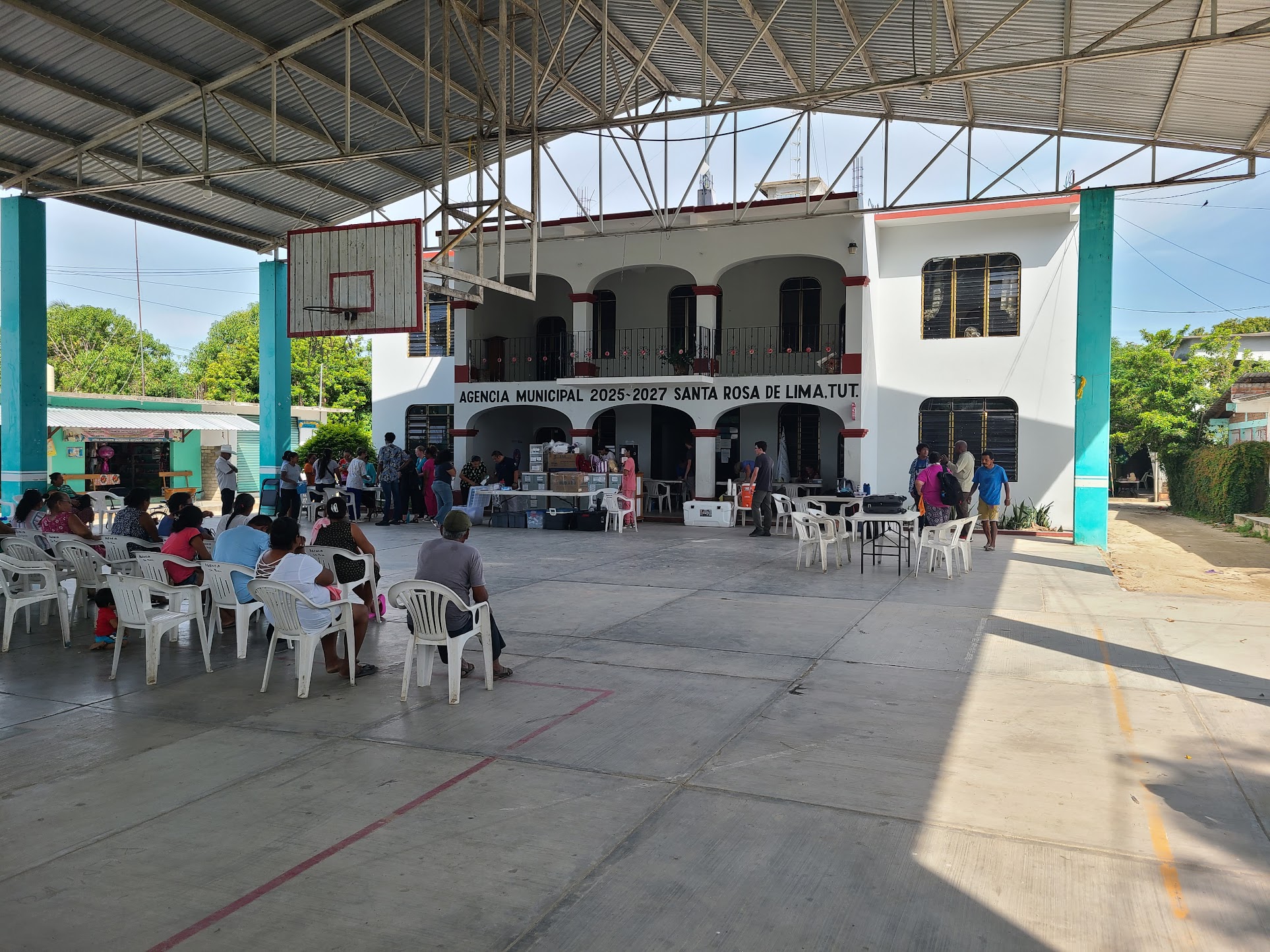 People sit in rows of white chairs under a large covered basketball court, waiting near tables set up for a community outreach event.