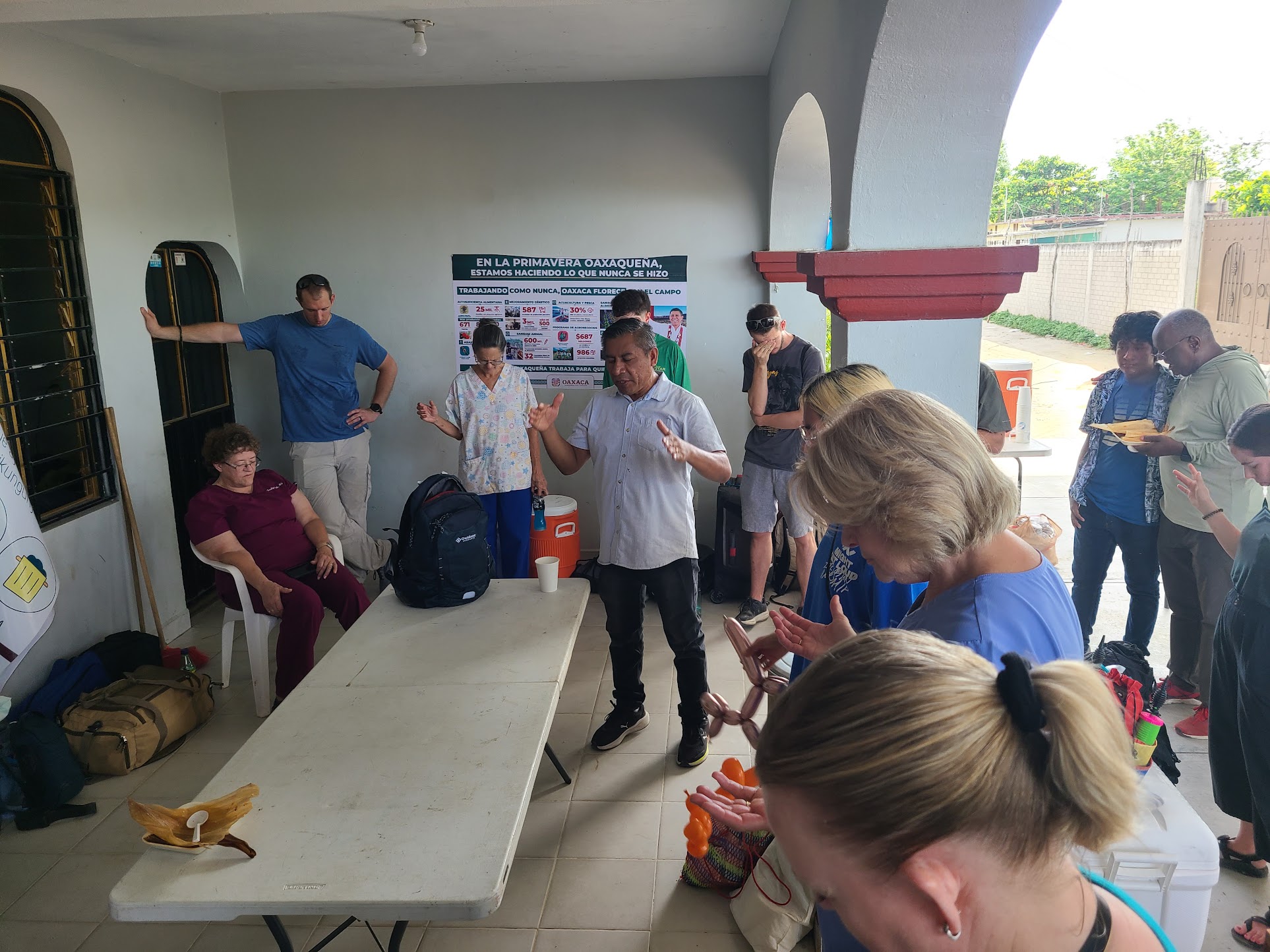A group of volunteers and locals stand and sit in prayer around a table under a shaded area outside a community building.