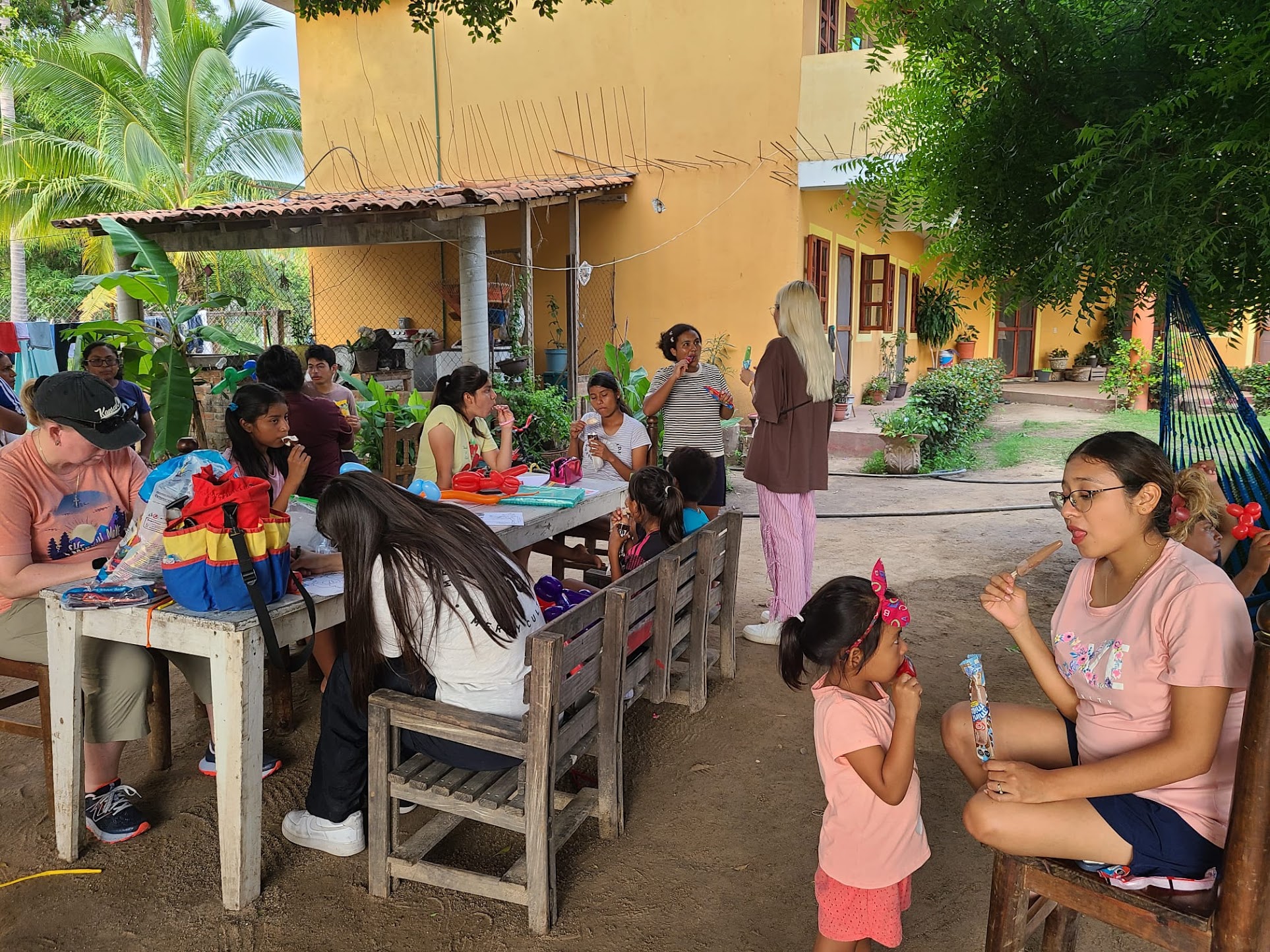 A group of children and adults gather around outdoor tables under shade trees, enjoying popsicles and crafts near a yellow building.