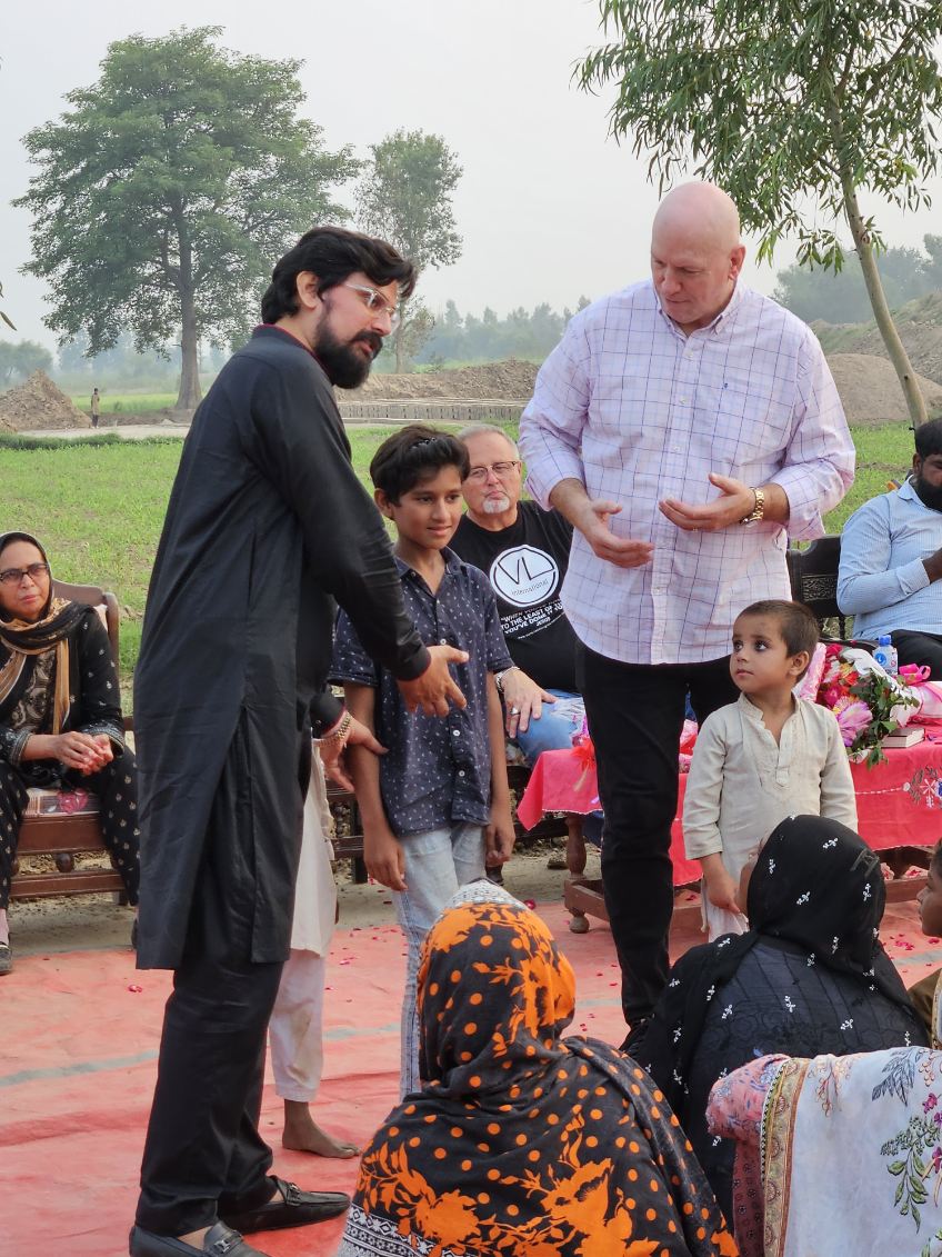 Pastor David speaks to a seated group of villagers outdoors while another interprets beside him during a rural outreach event.