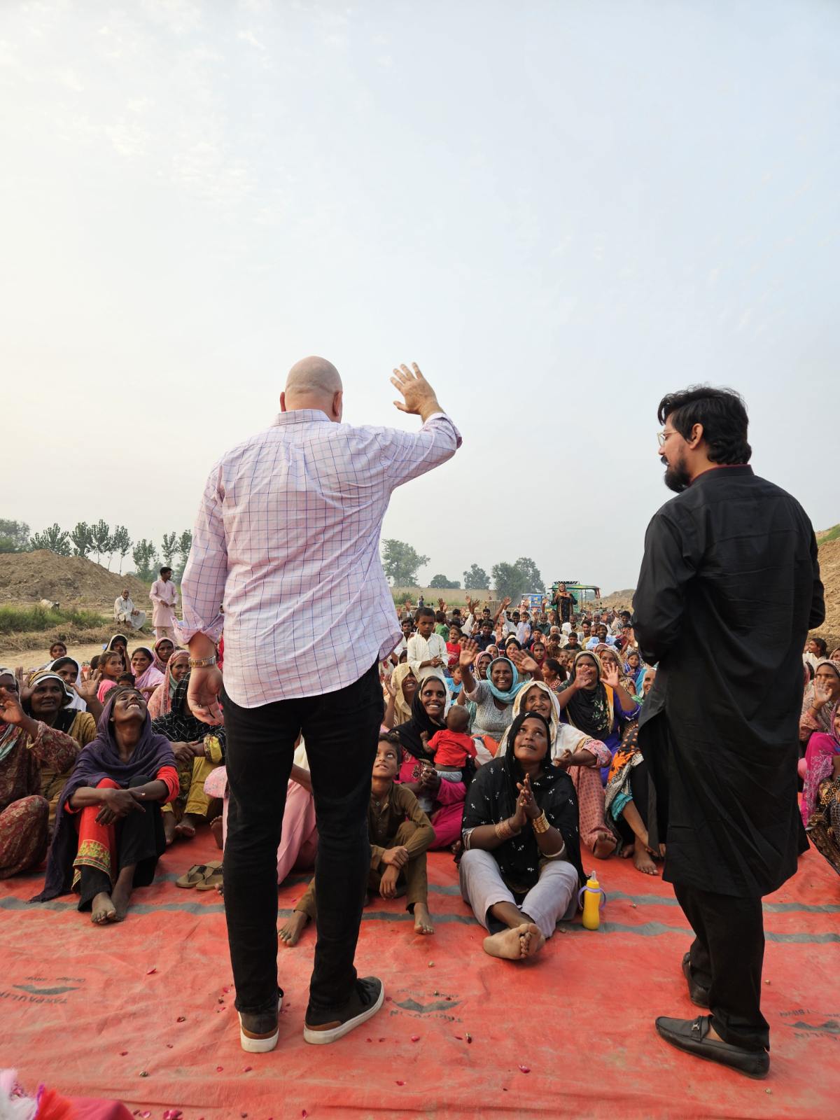 Pastor David stands before a crowd sitting on red cloth outdoors, addressing them during a daytime ministry meeting.