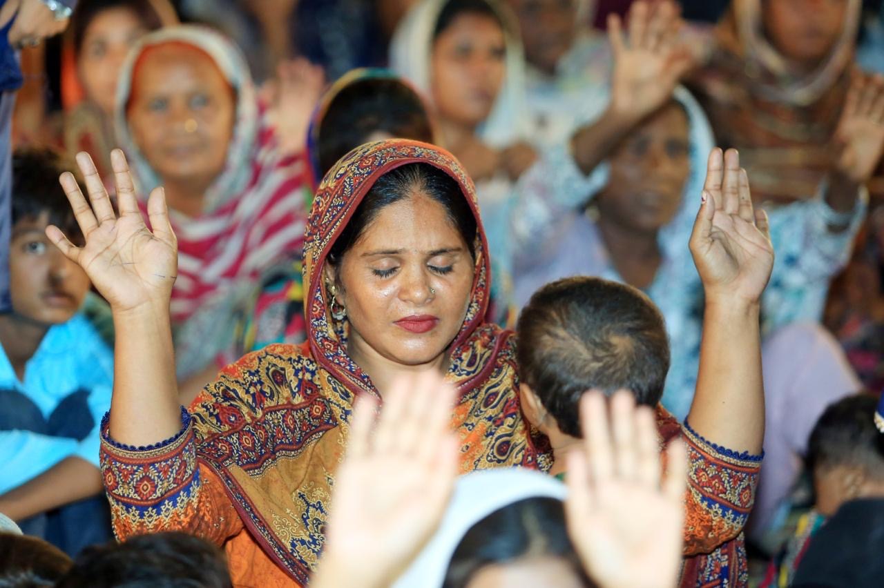 A woman in a colorful headscarf prays with eyes closed and hands raised among a large group of people during a worship service.