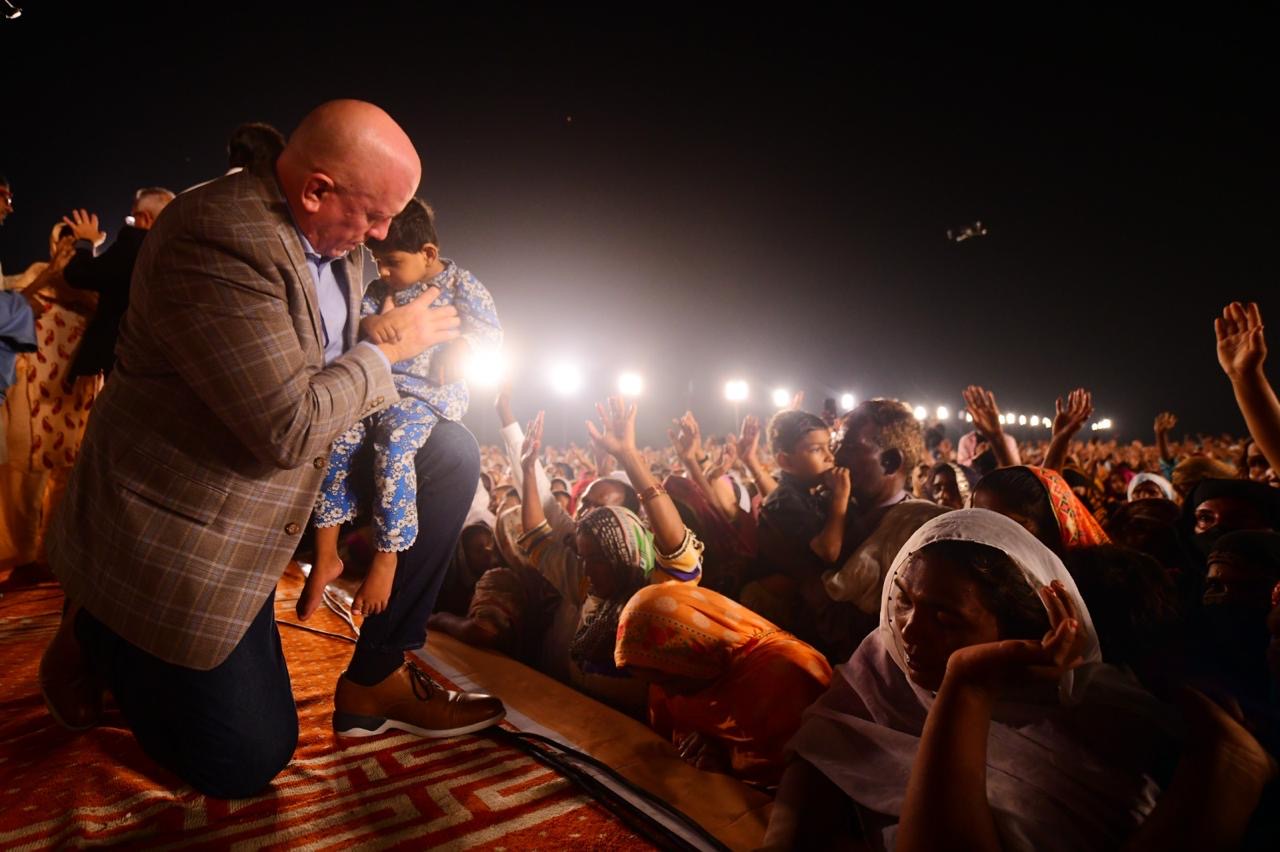 Pastor David kneeling in prayer with a child during a nighttime outdoor service in Pakistan, surrounded by a large crowd with raised hands.