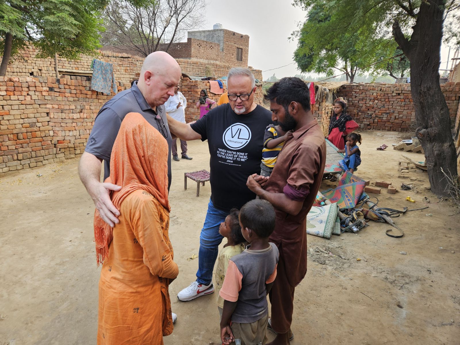 Pastor David and Ed pray with a family outdoors near a brick wall, surrounded by children and local villagers.