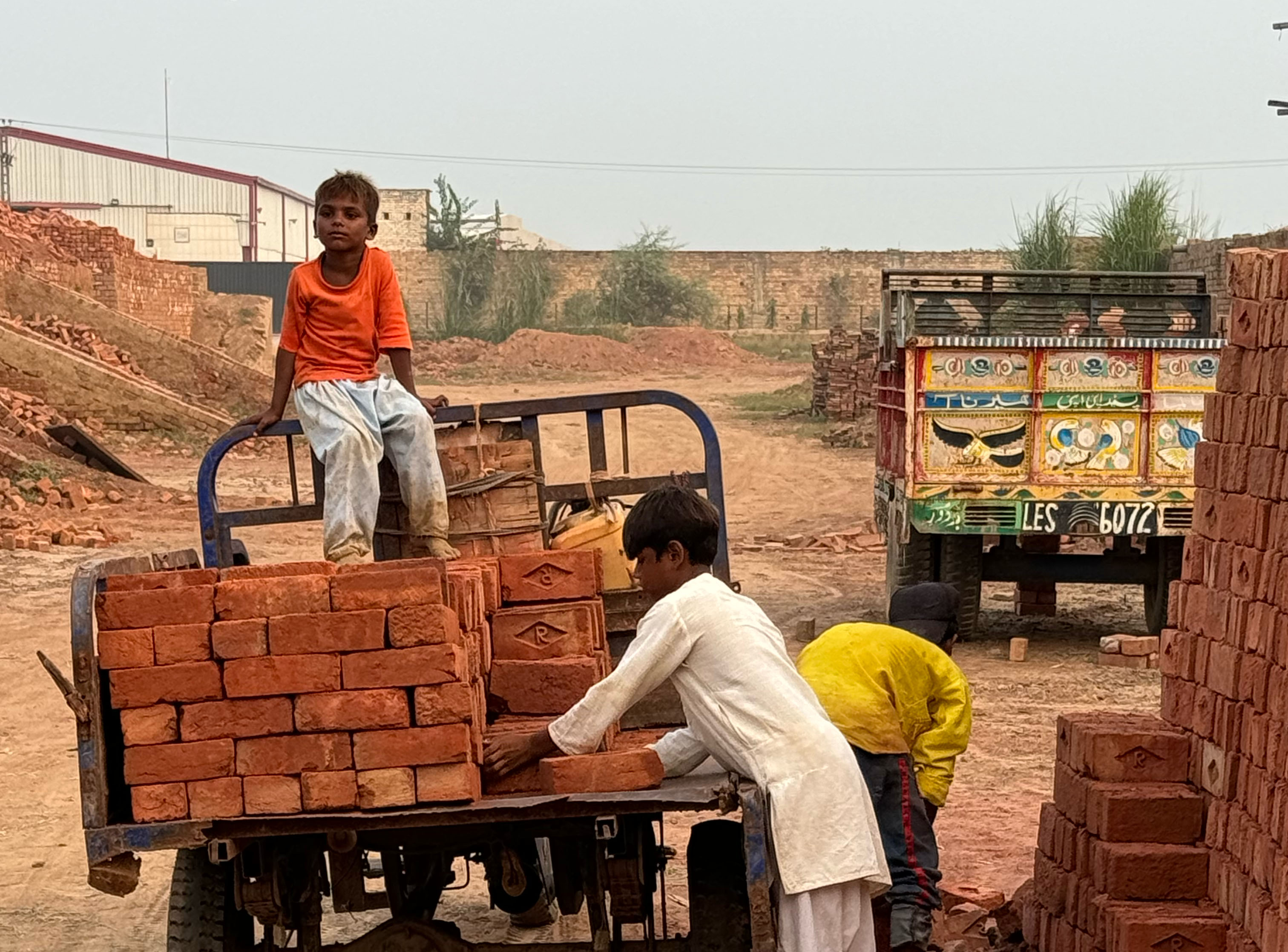 Young boys load red clay bricks onto a small truck at a brick kiln site, with piles of bricks and vehicles in the background.