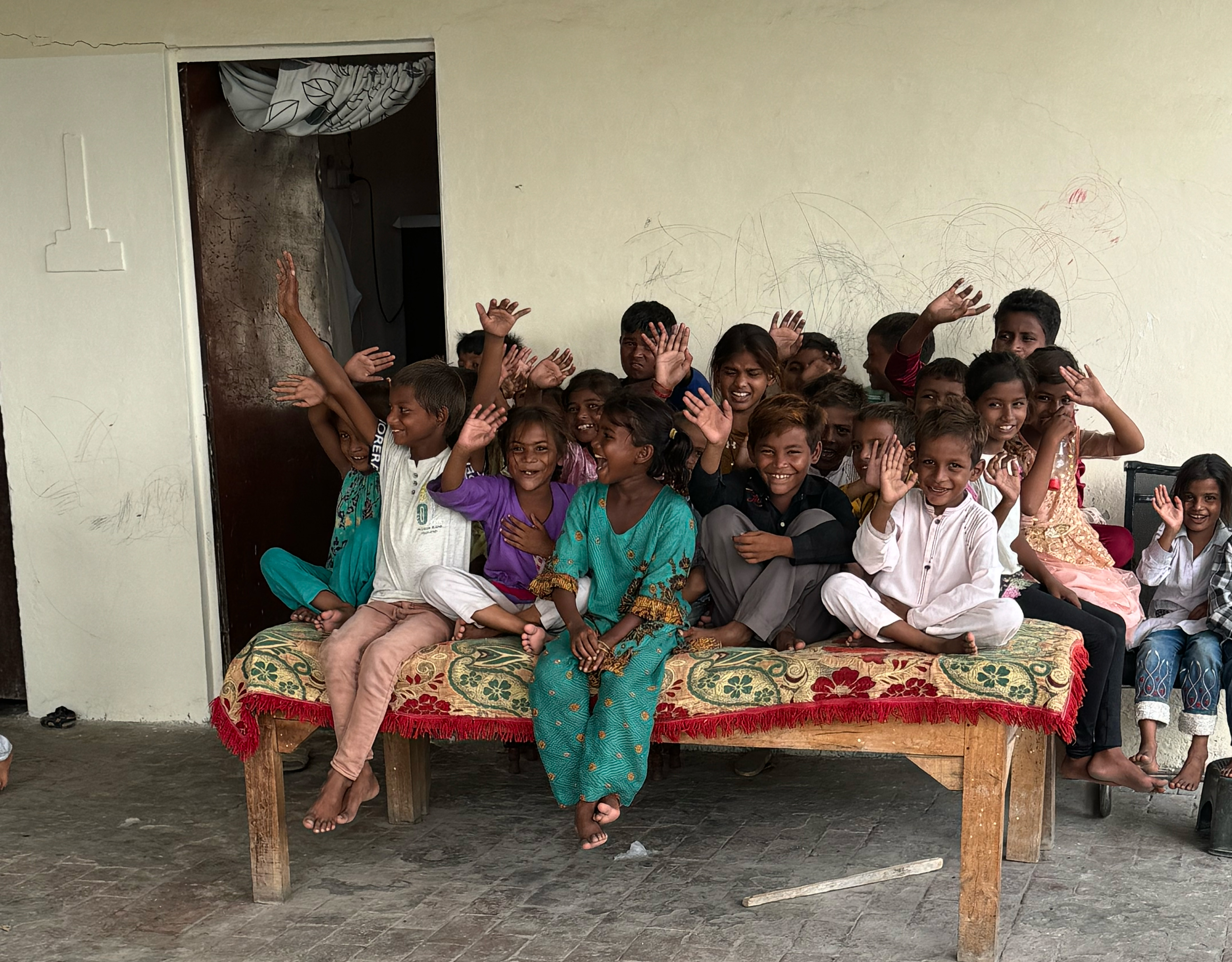 A cheerful group of children sit together on a wooden bed, waving and smiling inside a building with light-colored walls.