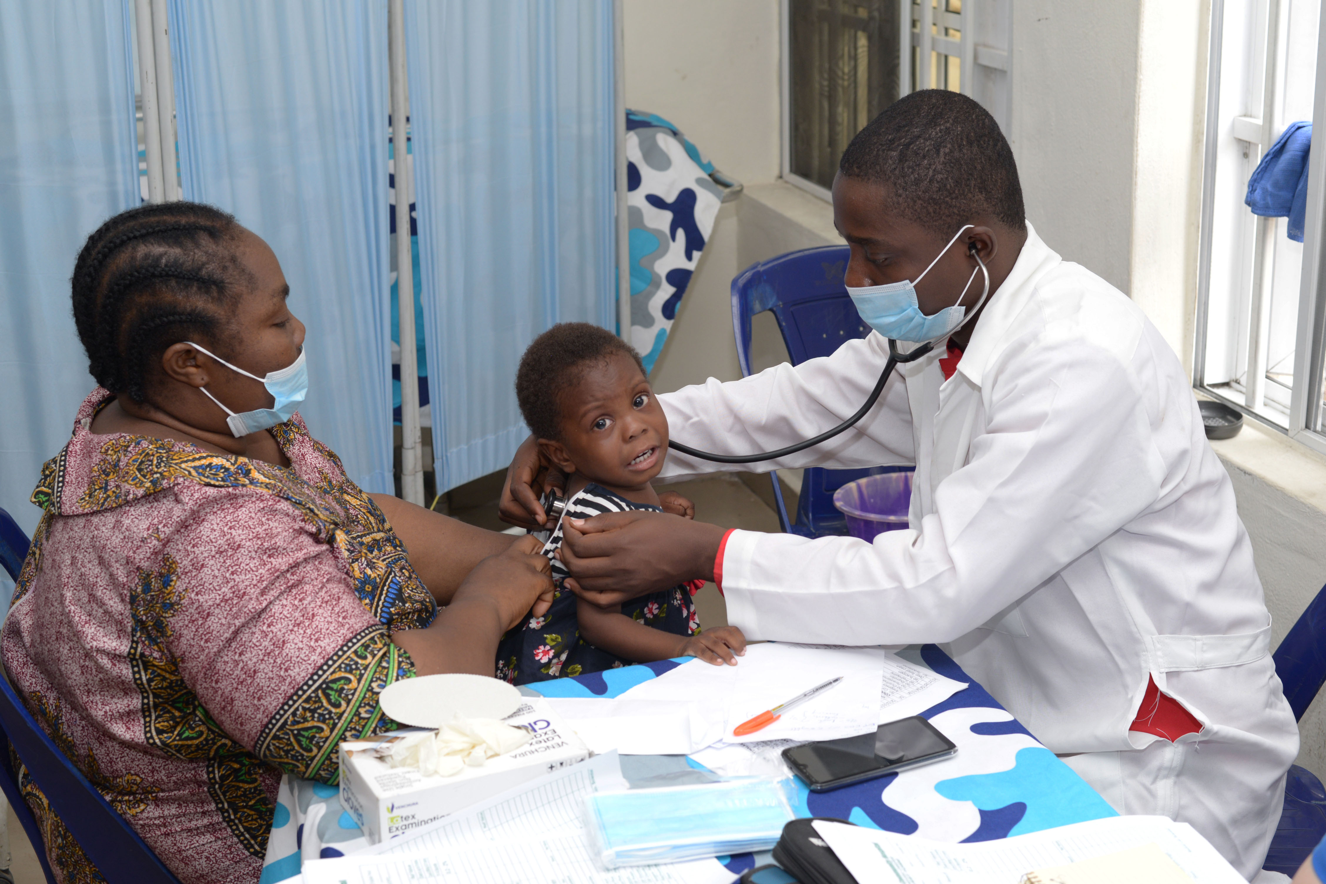 A doctor listens to a young child’s chest with a stethoscope while a caregiver looks on during a community health clinic visit.