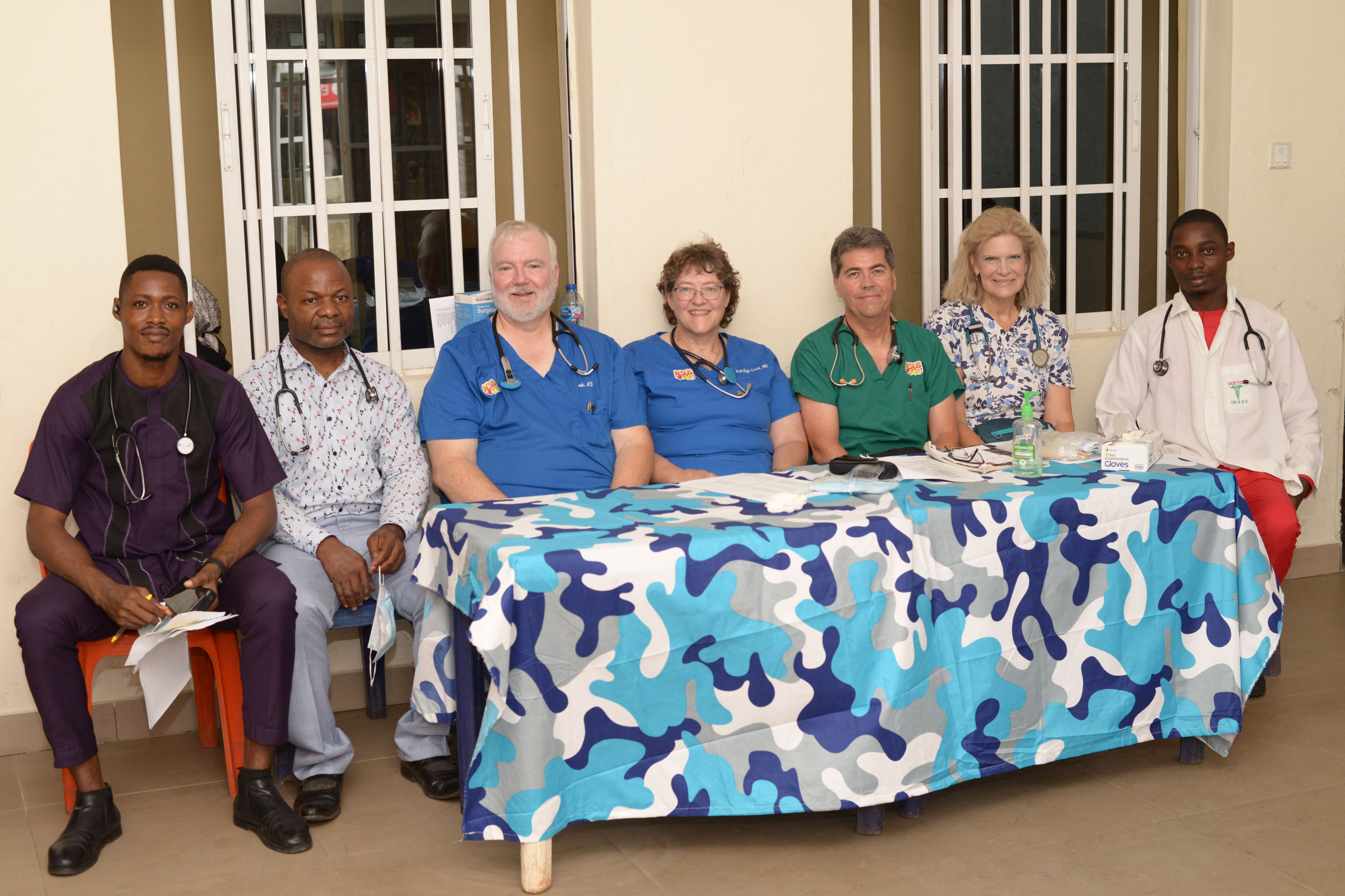 A group of healthcare professionals, both local and visiting, sit together behind a table covered with a blue patterned cloth inside a clinic.