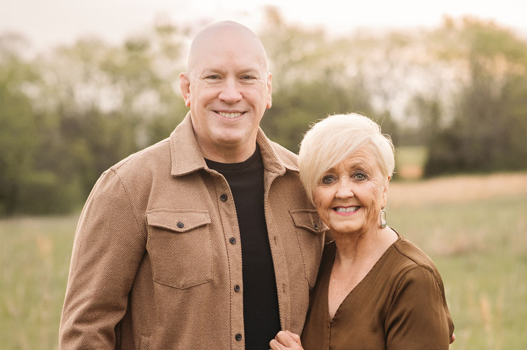 Smiling middle-aged man and woman standing outdoors in a field with trees in the background.