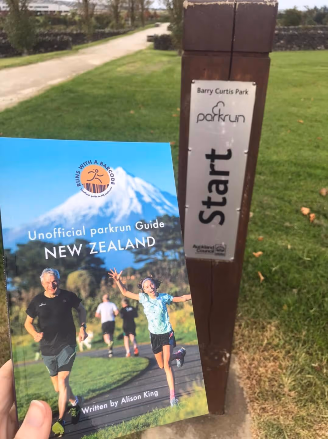 The cover of the runs with a bar code book is held up near a marking post for the start of a park run