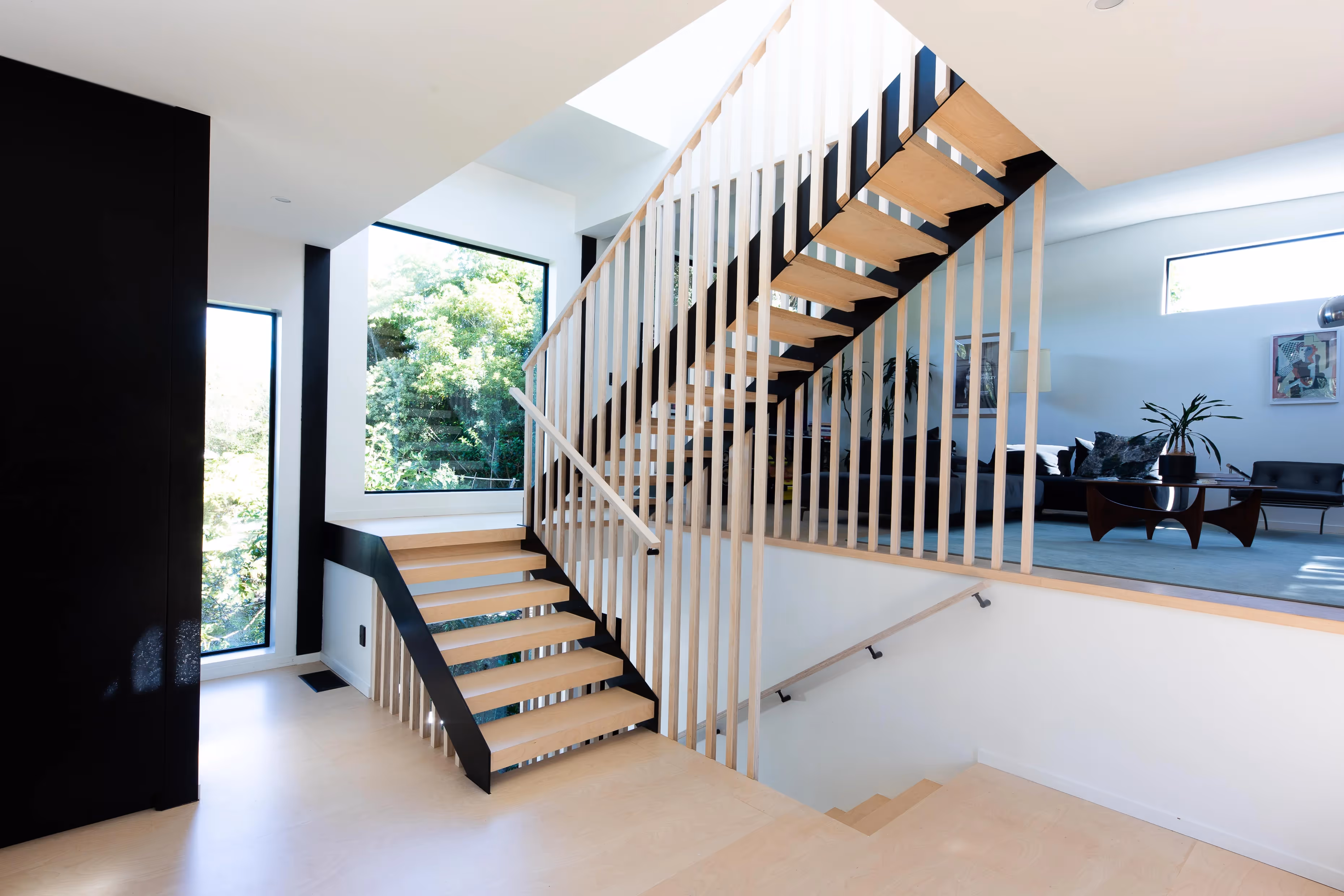 A modern, brightly lit interior features a wooden staircase with black handrails and vertical white balusters. The stairs lead to an upper level with large windows, white walls, and a glimpse of a stylish living area with contemporary furniture and artwork.