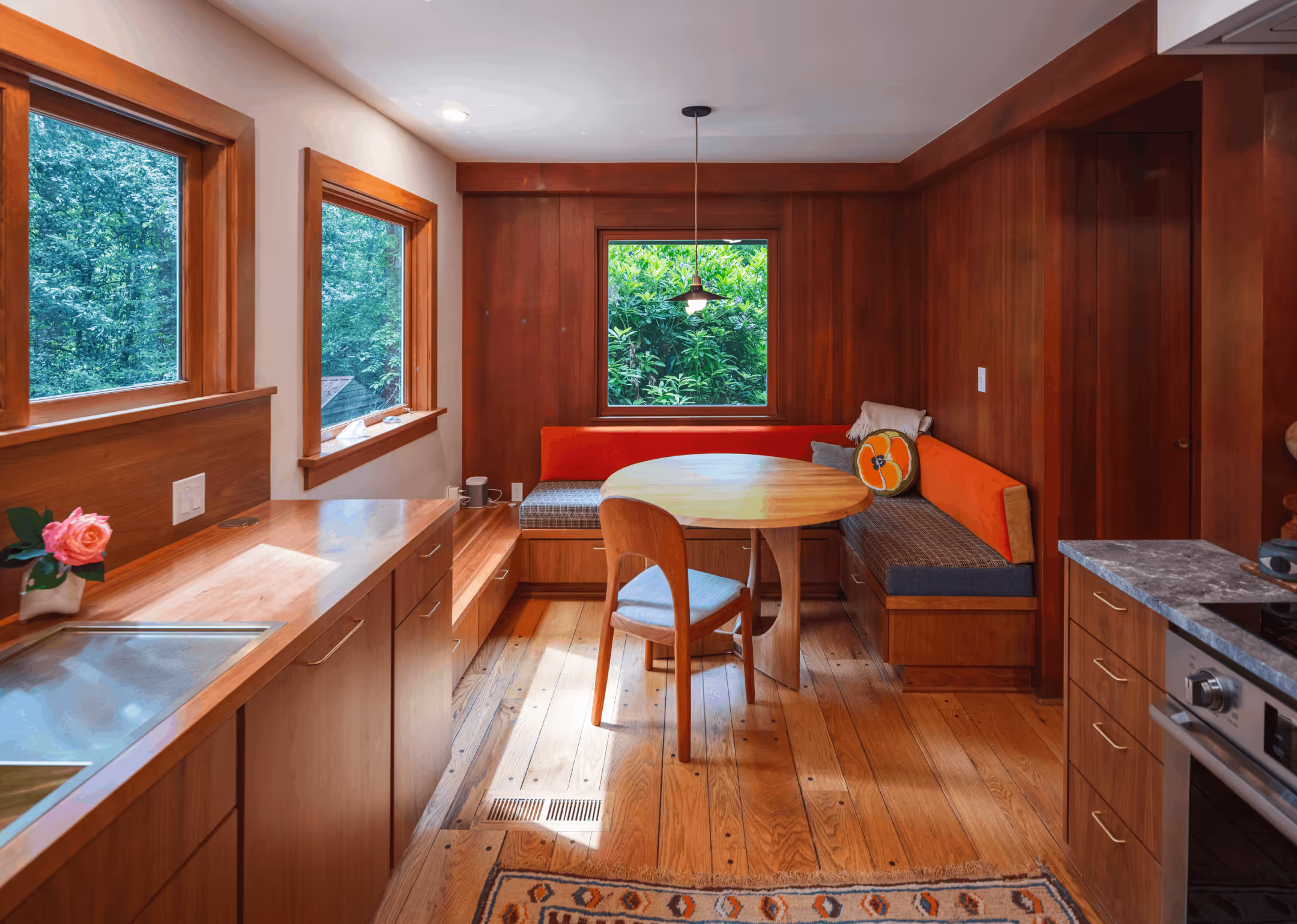Cozy wooden kitchen with built-in dining nook featuring orange cushions, round table, chair, and large windows with greenery outside.