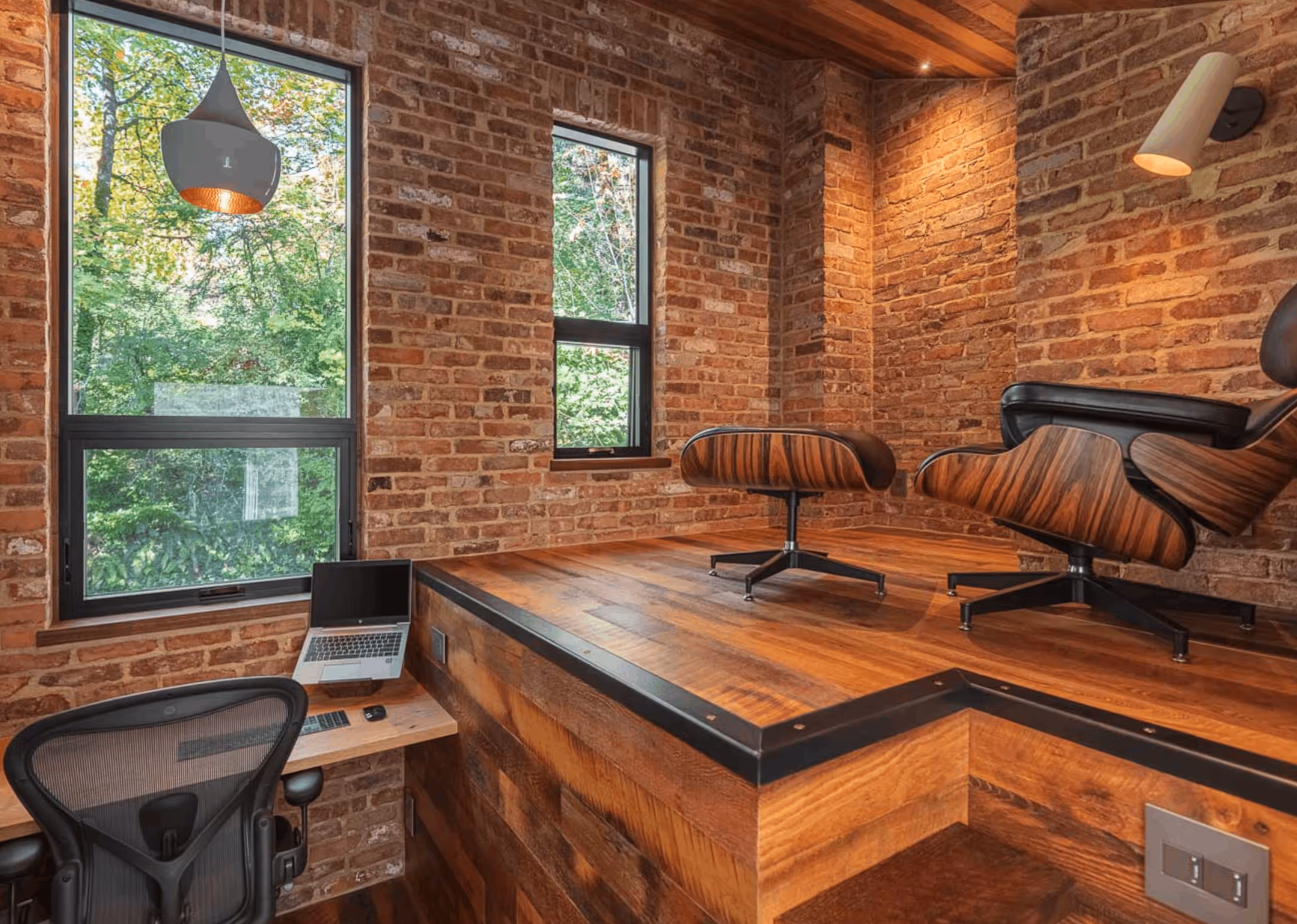 Cozy workspace featuring exposed brick walls, wood flooring, two mid-century modern chairs, a laptop on a floating desk, and large windows showing green trees outside.