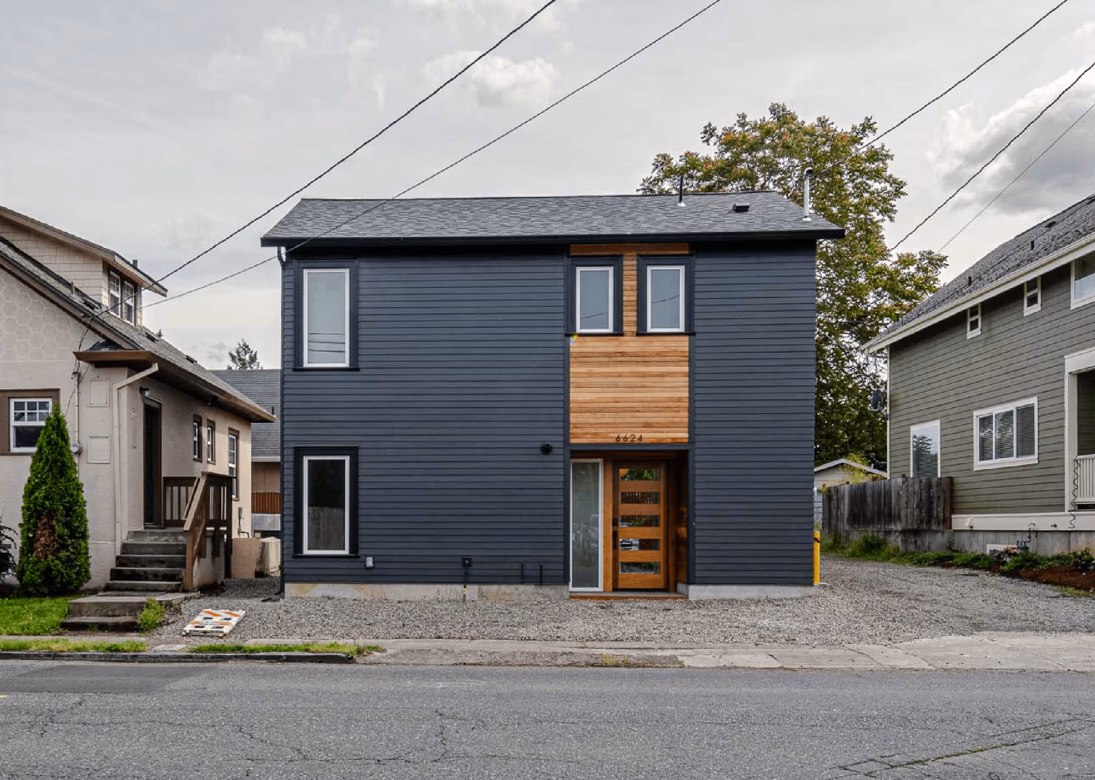 Modern two-story house with dark gray siding and wooden accents around the front door and windows, situated between two older homes.