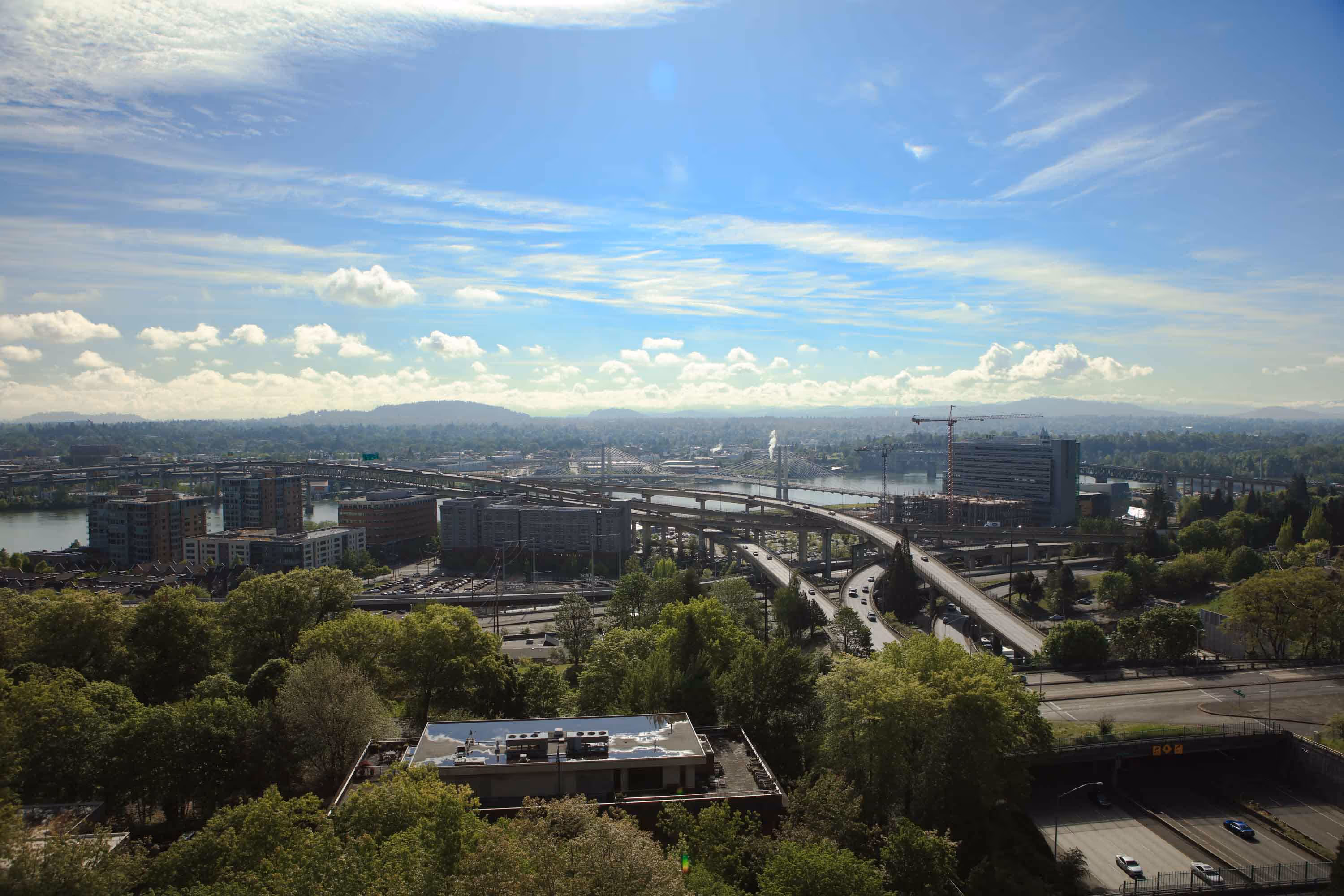 Cityscape with highways, bridges over a river, mid-rise buildings, and green trees under a partly cloudy blue sky.