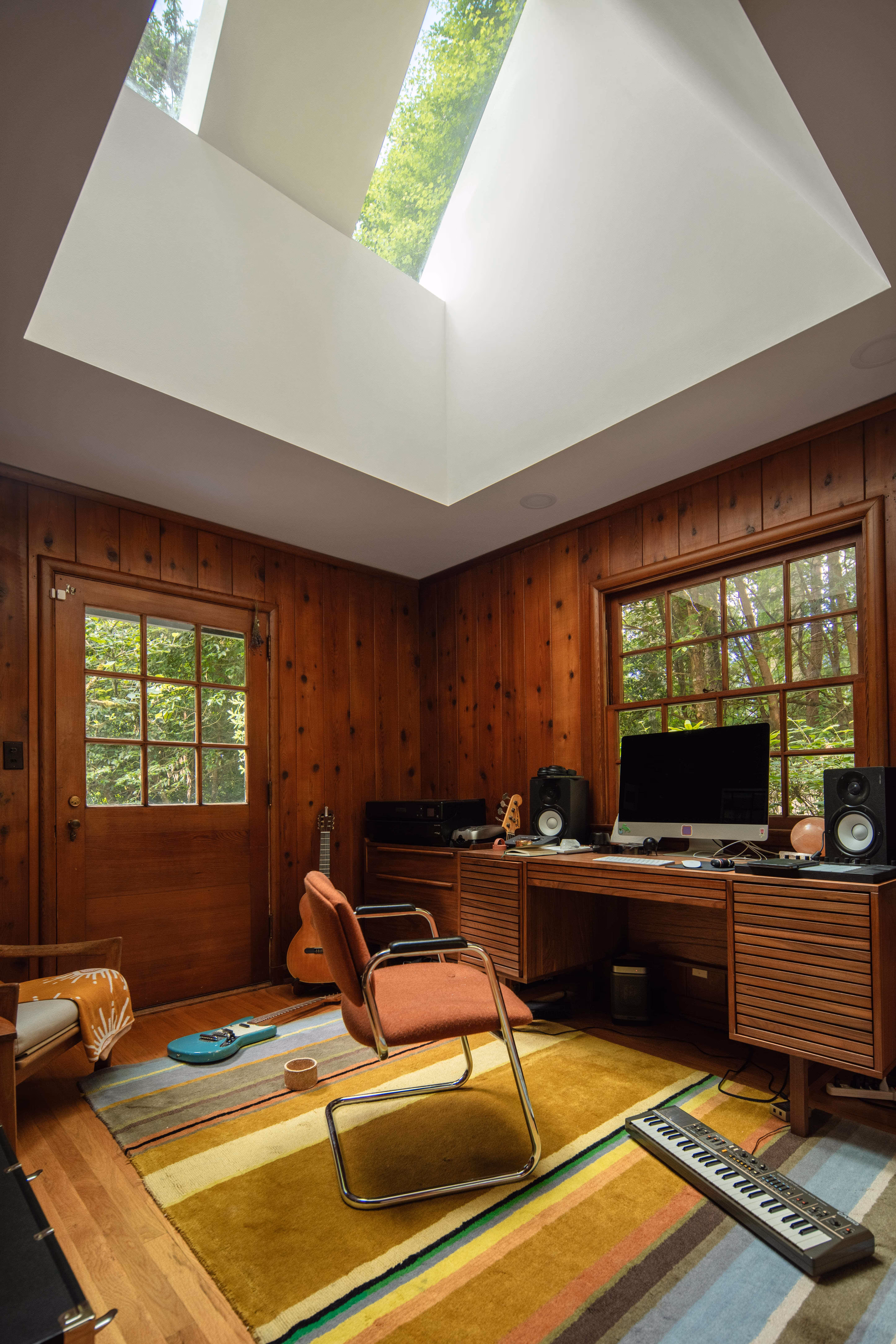 Home office with wood-paneled walls, skylight, wooden desk with computer and speakers, orange chair, guitar, and keyboard on multi-colored rug.