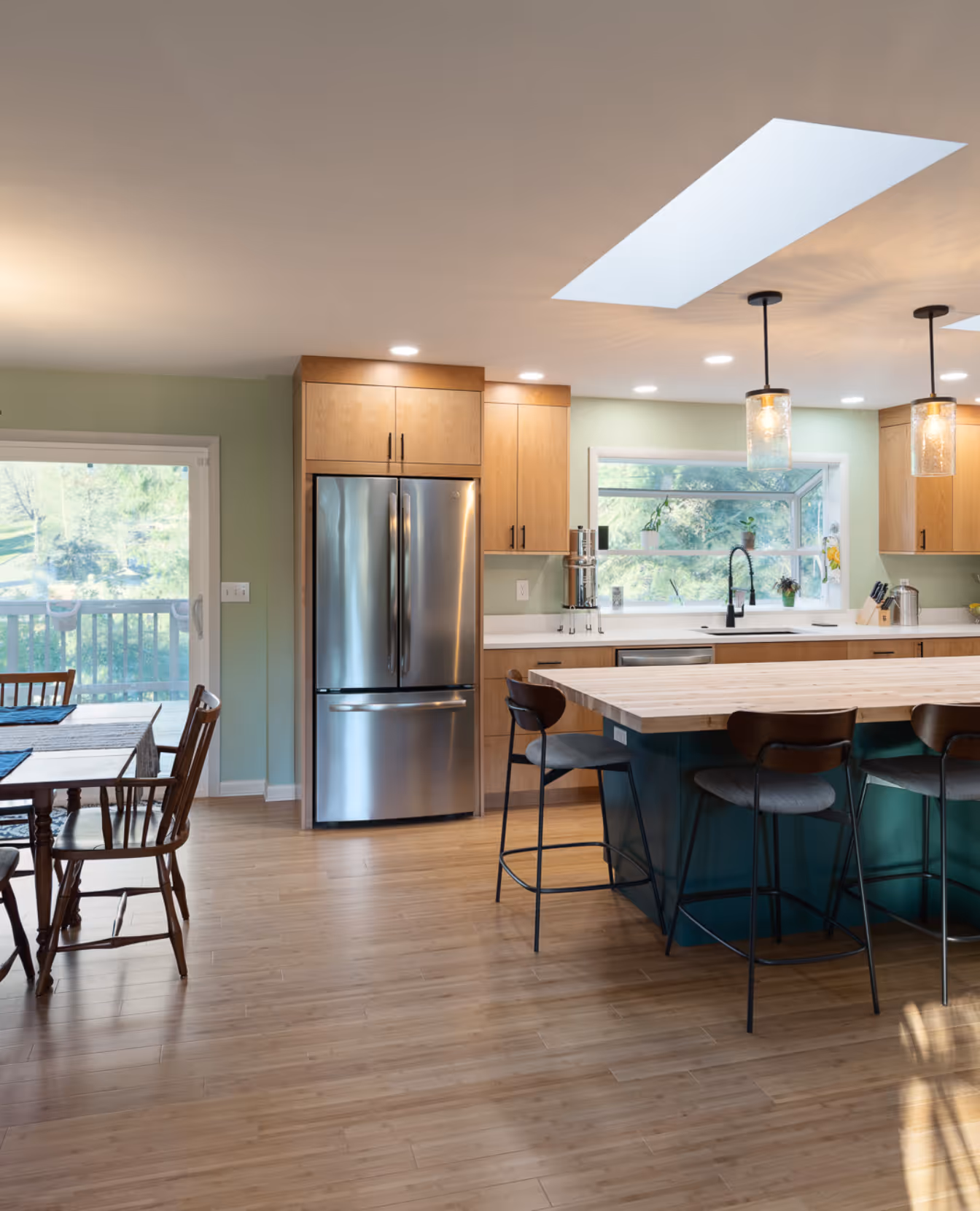 Modern kitchen with wooden cabinets, a stainless steel refrigerator, an island with bar stools, pendant lights, and a dining table near sliding glass door.