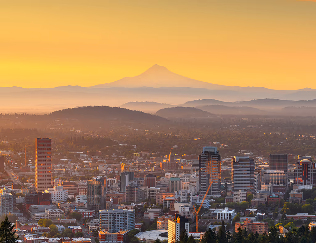 City skyline at sunset with buildings in foreground and mountains with a prominent peak in the background.