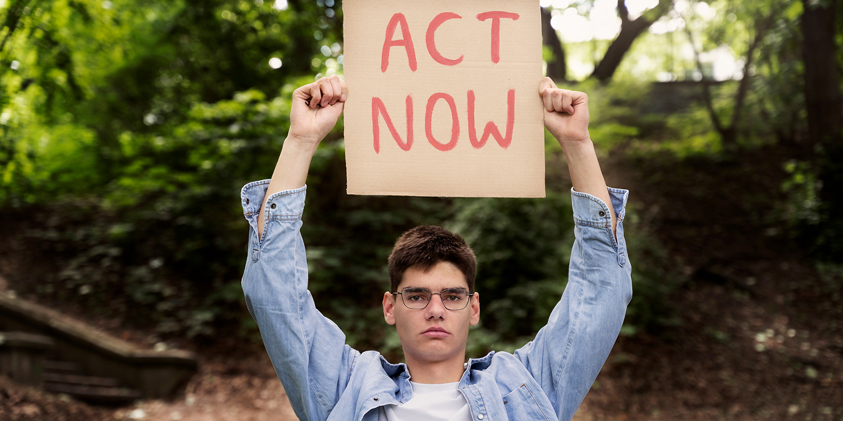 Young man in glasses and denim jacket holding a cardboard sign that says 'ACT NOW' in a forested area.