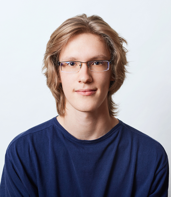 Young person with light brown hair and glasses wearing a navy blue shirt, facing forward against a plain light background.
