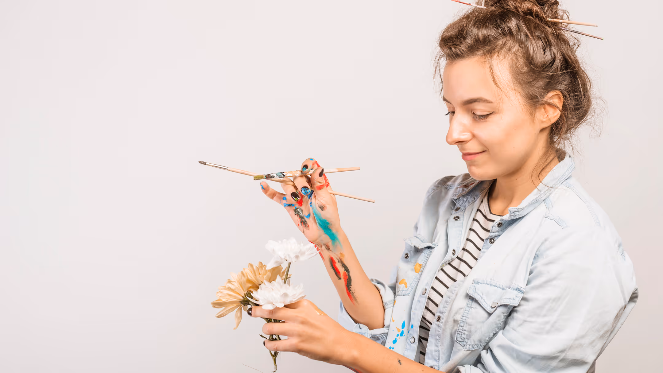 Young woman with paint on her hands and arms holding paintbrushes and flowers, smiling slightly.