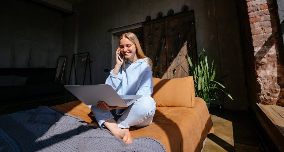 Mensch sitzt auf Bett mit Telefon in der Hand und Laptop auf dem Schoß