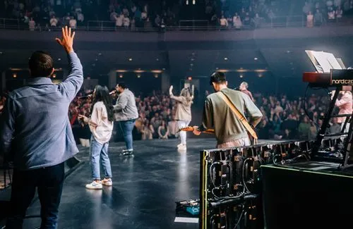 Musicians and singers performing on stage in front of a large audience with one person raising a hand.