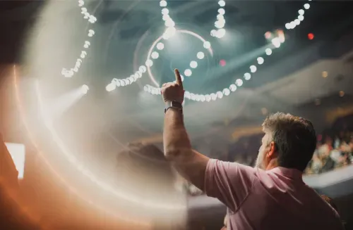 Man in a pink shirt raising his hand with thumb up at an indoor event with bright circular lights overhead.