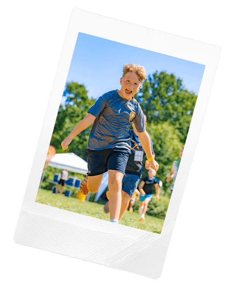 Smiling boy wearing a maroon Jefferson Elementary t-shirt and a lanyard, standing outdoors on a sandy ground with arms outstretched.