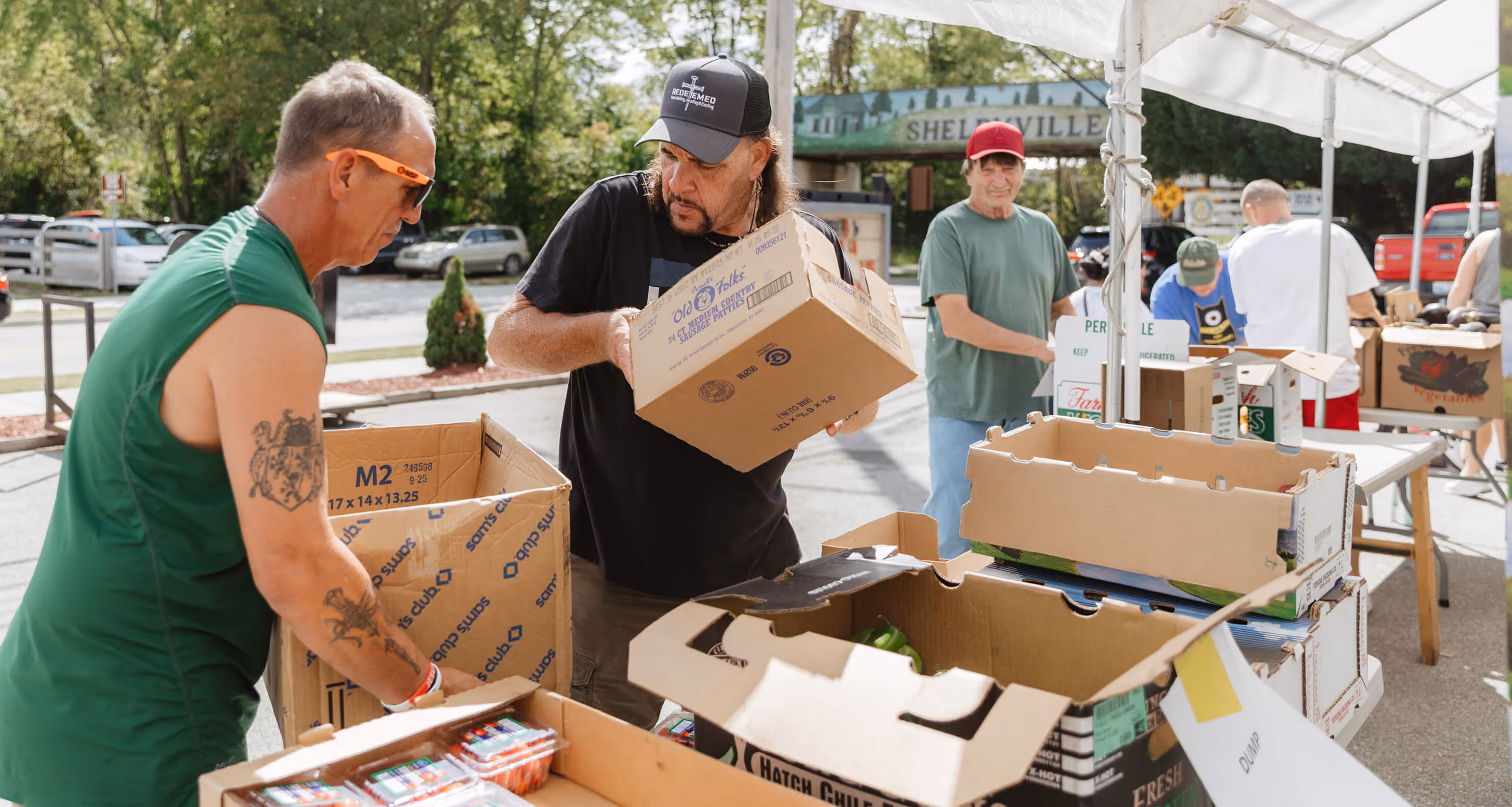 Volunteers sorting and packing food items under a white tent at an outdoor community food distribution event.