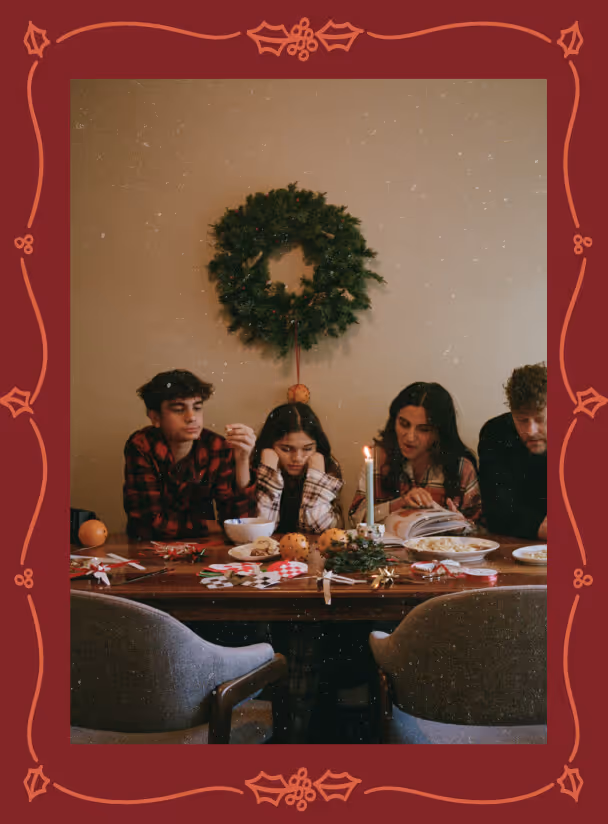 Four people sitting at a wooden table decorated with holiday items, including a lit candle and greenery, with a wreath hanging on the wall behind them.