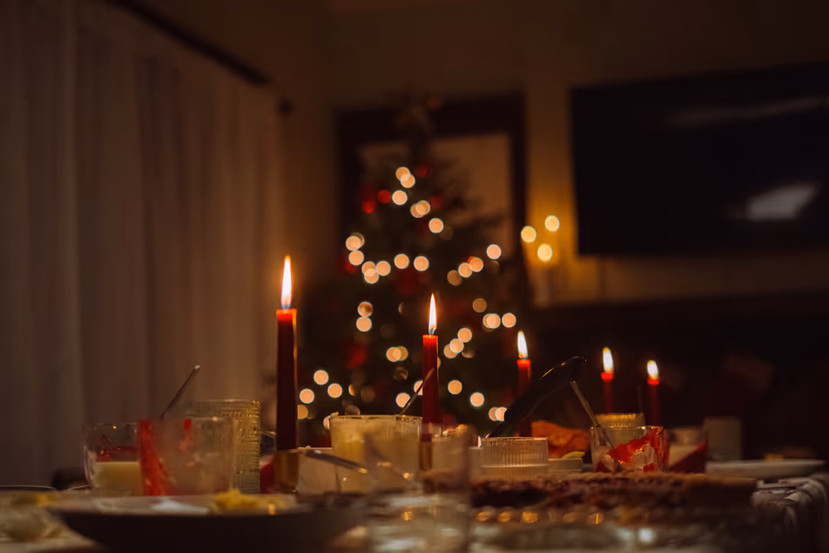 Dimly lit dining table with lit red candles and a blurred decorated Christmas tree in the background.