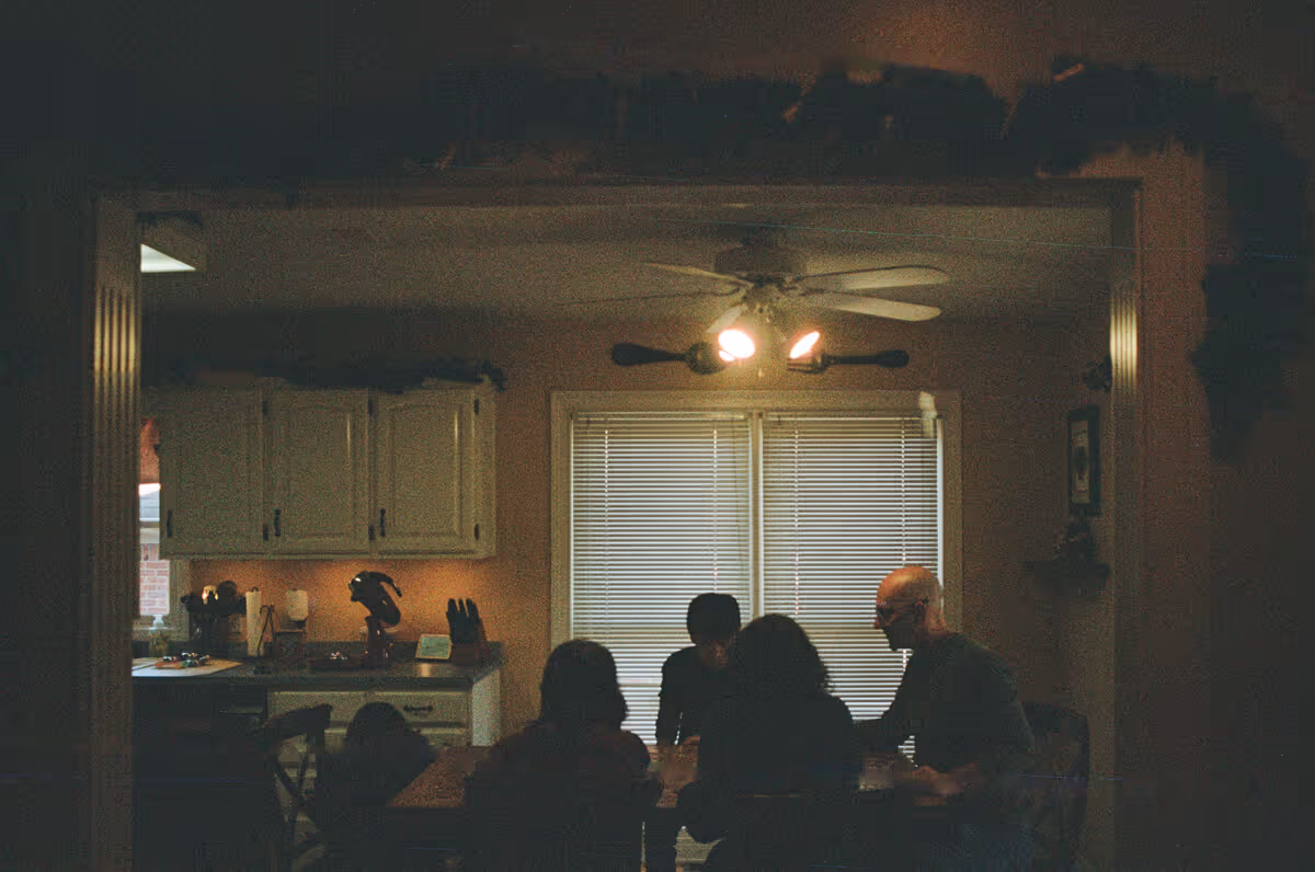 Silhouettes of four people sitting around a dining table in a dimly lit kitchen with closed blinds and ceiling fan lights turned on.