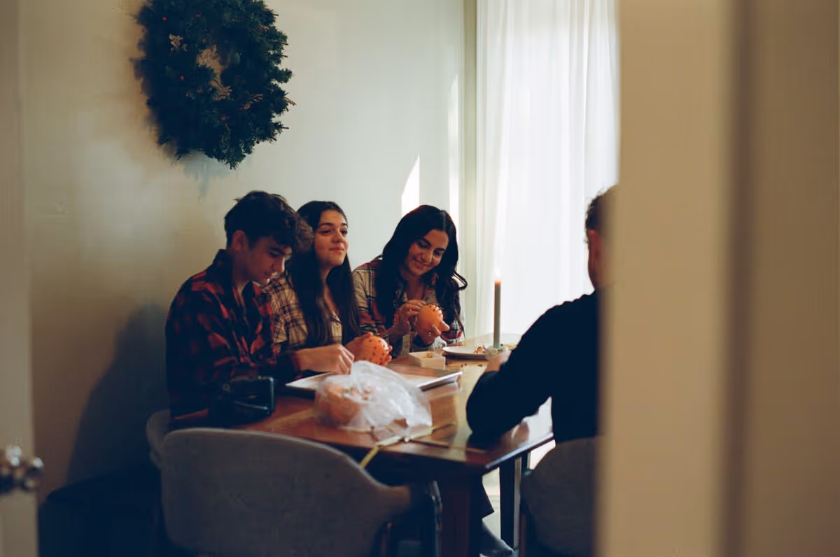 Four people sitting at a dining table engaged in decorating orange crafts, with a Christmas wreath on the wall in the background.