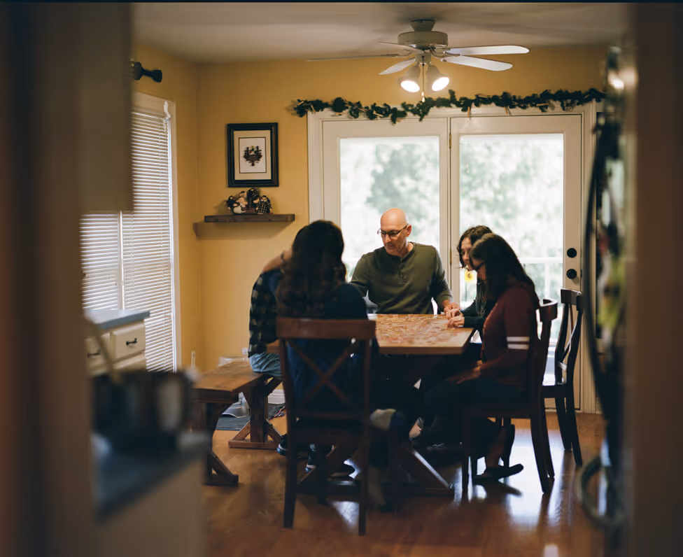Family of five holding hands and praying around a dining table in a warmly lit home.