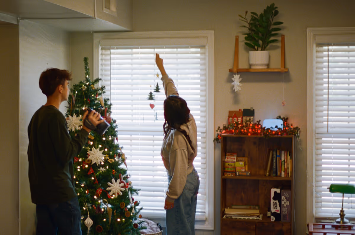 Two people decorating a Christmas tree in a cozy room with shelves and window blinds.
