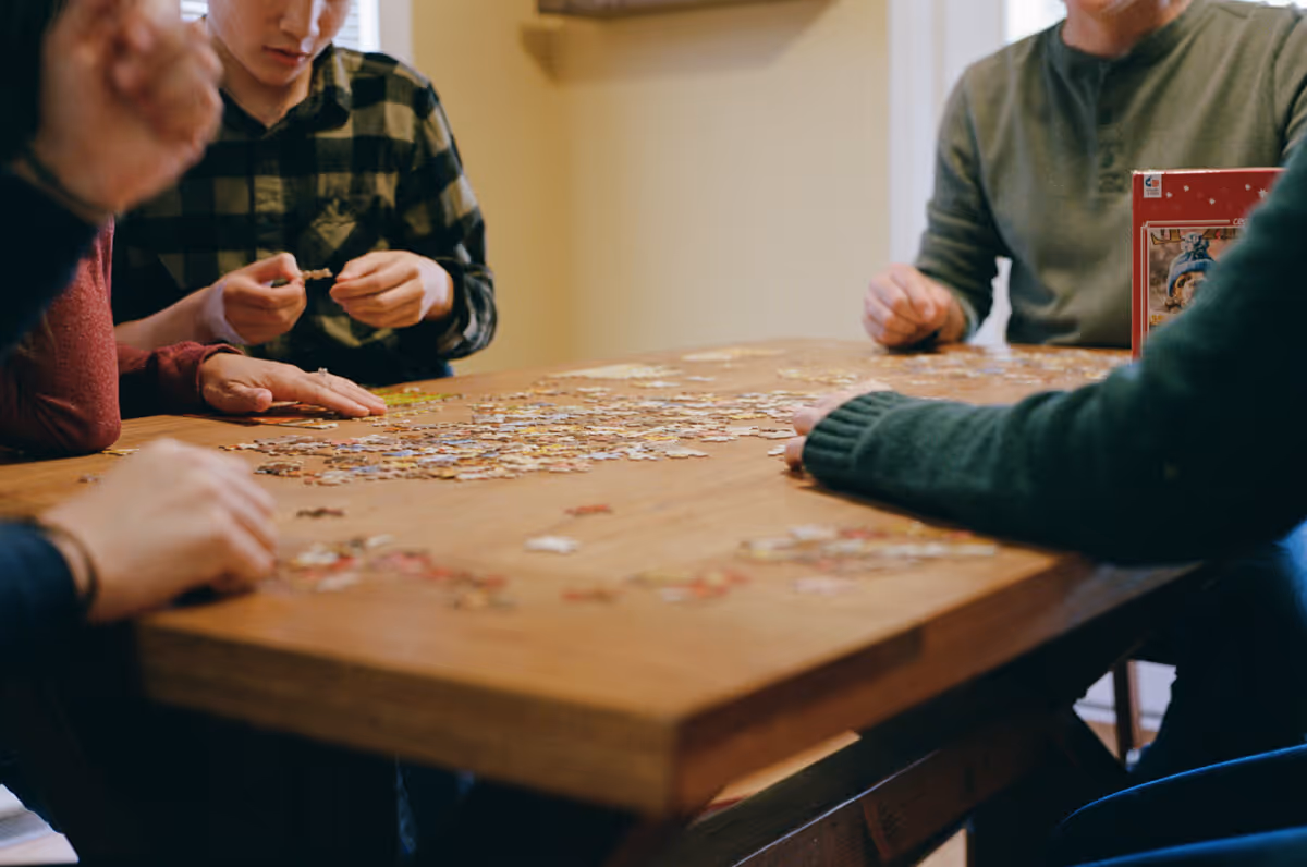 Four people sitting around a wooden table assembling a jigsaw puzzle in a cozy room.