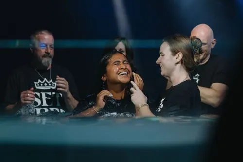 A woman smiling joyfully while being baptized in water, surrounded by three people assisting her.