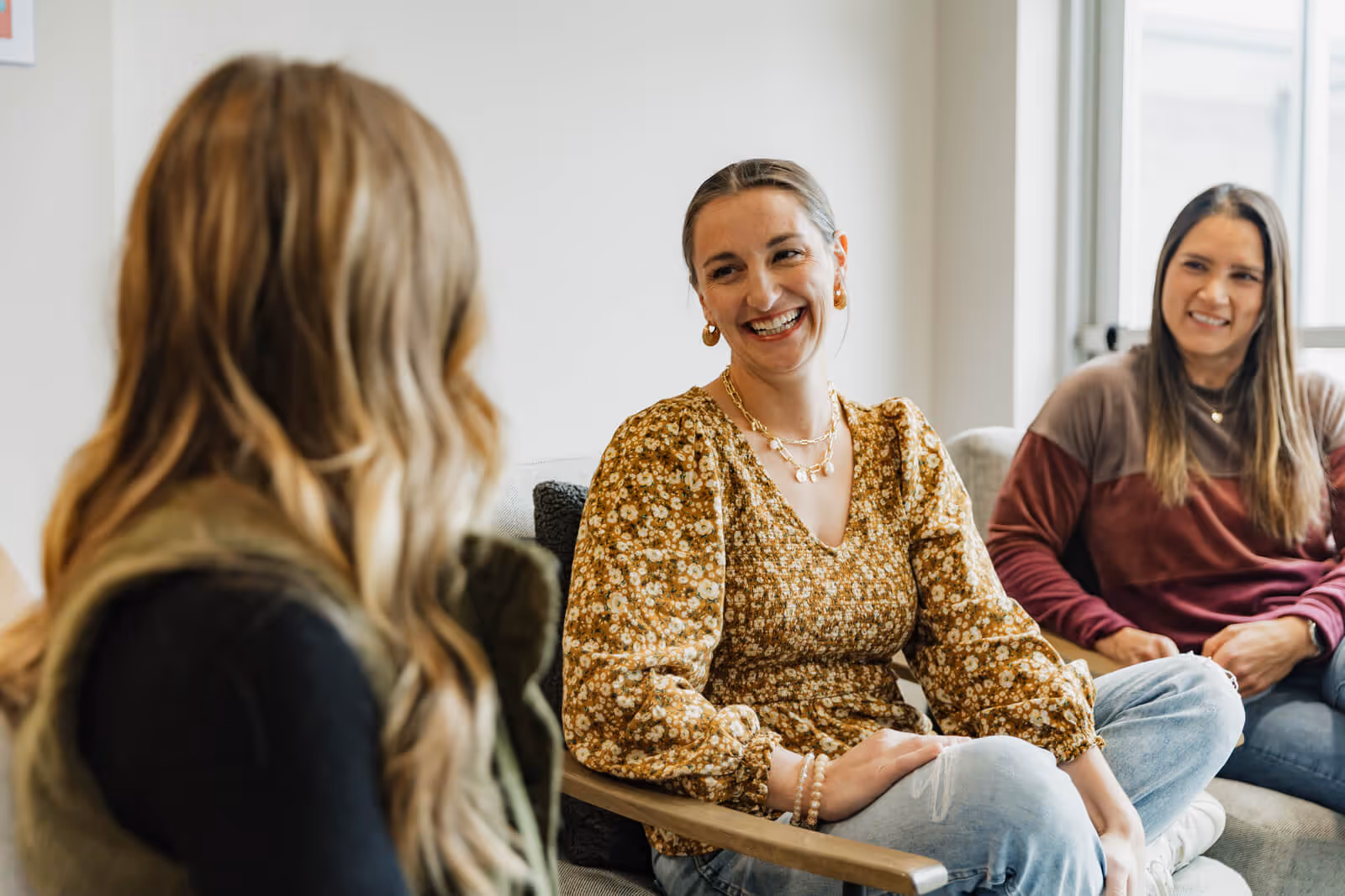 Three women sitting indoors on a couch and chair, smiling and engaged in conversation.