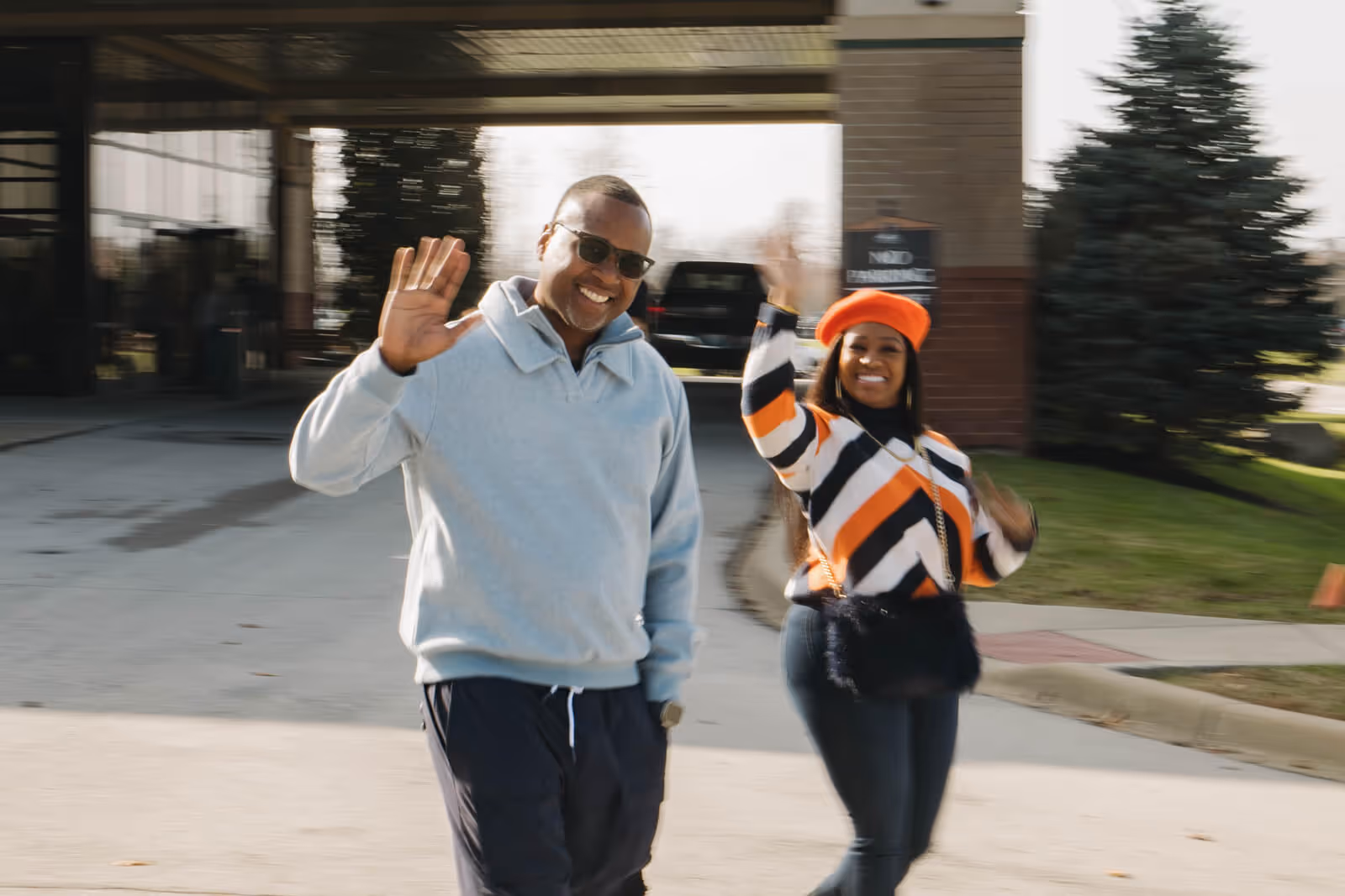 Man and woman smiling and waving while walking outside near a building and trees.