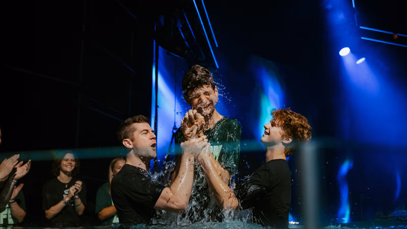 Three young men in water, two supporting a third man who is being baptized with water splashing around them.