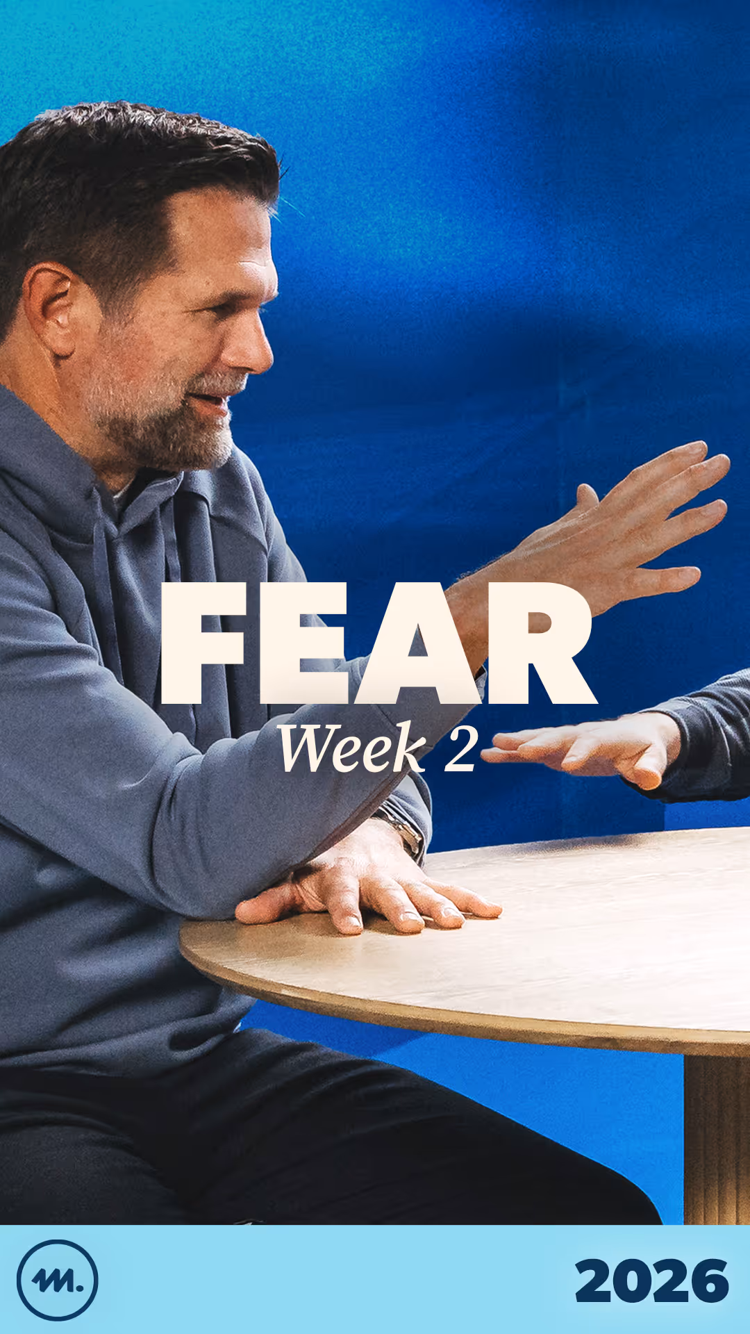 Man in gray hoodie reaching across a wooden table with 'FEAR Week 2' text overlay on a blue background.