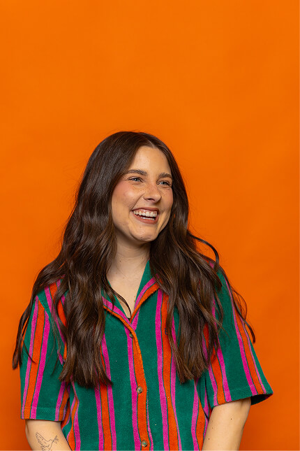 Smiling woman with long wavy brown hair wearing a green shirt with red and pink stripes against an orange background.